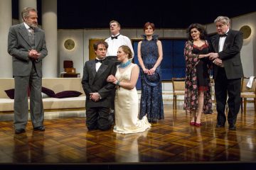 Lavalla on seitsemän juhlapukuista näyttelijää, joista kaksi polvistuu keskellä. Muut seisovat heidän ympärillään ja näyttävät olevan mukana dramaattisessa hetkessä. Lavastus muistuttaa ylellistä olohuonetta, jossa on tyylikäs kalustus.