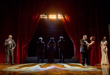 Punaisilla verhoilla ja dramaattisella valaistuksella varustetulla teatterilavalla näkyy oikealla kolme ihmistä, mukaan lukien punapukuinen nainen, jotka ovat vuorovaikutuksessa, vasemmalla yksi mies, joka seisoo yksin, ja neljä siluettihahmoa, jotka seisovat kirkkaisiin valoihin päin keskellä lavaa.