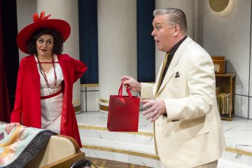 Punaiseen hattuun ja takkiin pukeutunut nainen seisoo kermanväriseen pukuun pukeutuneen miehen vieressä, joka pitää kädessään punaista lahjapussia ja näyttää puhuvan vilkkaasti. He ovat näyttämöllä, jonka taustalla on valkoisia pylväitä.