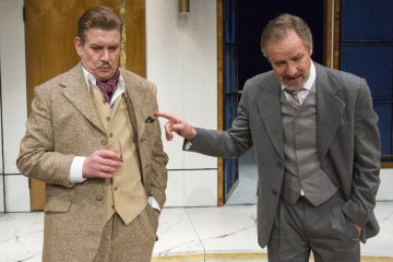 Kaksi vanhaan pukuun pukeutunutta miestä seisoo sisätiloissa; toisella on juoma kädessään ja katsoo mietteliäästi alaspäin, kun taas toinen tekee eleitä ja puhuu hänelle vaikuttaen vakuuttavalta tai painokkaalta.