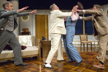 Neljä pukuihin pukeutunutta miestä on dramaattisessa kohtauksessa näyttämöllä, joka muistuttaa olohuoneen lavastusta; yksi valkoisiin pukeutunut mies tarttuu toista miestä kauluksesta, kun kaksi muuta miestä kurottautuvat väliin.