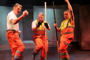 Kolme oransseihin työhousuihin ja heijastinliiveihin pukeutunutta miestä poseeraa innokkaasti lavalla pitelemässä pitkiä sauvoja ja tekemässä energisiä eleitä, aivan kuin he juhlisivat tai esiintyisivät teatteriesityksessä.