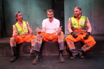 Kolme oransseihin työhousuihin ja saappaisiin pukeutunutta miestä istuu betonipenkillä vesipullot kädessään. Keskimmäisellä miehellä on valkoinen paita, kahdella muulla heijastinliivit ja hanskat, ja he näyttävät puhuvan ja nauravan yhdessä.