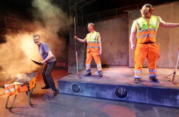 Lavalla esiintyy kolme miestä; kaksi oransseihin rakennuspukuihin pukeutunutta miestä seisoo lavalla pitelemässä työkaluja, kun taas toinen, rennosti pukeutunut mies on vuorovaikutuksessa etualalla olevan savuavan kottikärryn kanssa. Lavavalot ja savu luovat dramaattisen tunnelman.