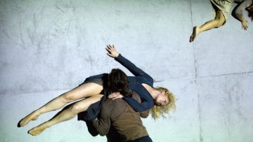 Kaksi sinisiin vaatteisiin pukeutunutta ihmistä näyttää leijuvan tai putoavan harmaalla pinnalla, ja kolmannen henkilön jalat näkyvät oikeassa yläkulmassa. Heidän kätensä ja jalkansa ovat ojennettuina, mikä viittaa liikkeeseen tai tanssiin.