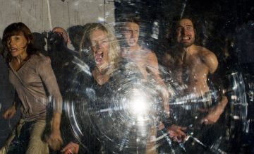 Neljä ihmistä, joista osa on paidattomia, näyttävät huutavan tai olevan hädissään säröisen, pyöreän särkyneen lasin takana, jonka keskellä heijastuu kirkas valo, mikä luo dramaattisen ja kaoottisen kohtauksen.