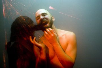 Paidaton, parrakas henkilö seisoo dramaattisessa valaistuksessa, suu auki ilmeikkäässä asennossa. Hänen viereensä ilmestyy tumma, varjoinen heijastus tai hahmo, joka luo surrealistisen, tunteikkaan tunnelman.