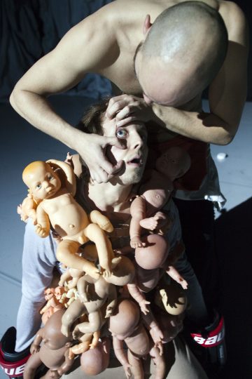 Kaksi ihmistä on dramaattisessa kohtauksessa; toinen pitää toisen silmää auki, kun istuva henkilö on peitetty muovisilla vauvanukeilla ja nuken osilla. Valaistus on kova, mikä luo surrealistisen ja intensiivisen tunnelman.