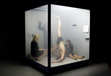 Kolme ihmistä istuu lasikuution sisällä, ja yksi heistä tekee käsilläseisontaa kahden muun katsoessa. Kohtaus on hämärästi valaistu, tunnelma on sumuinen ja kuution ulkopuolella seinällä on papereita.