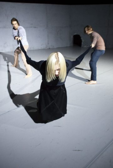 Nainen, jolla on pitkät vaaleat hiukset, polvistuu lattialle, kädet ojennettuina ja kahden hänen takanaan seisovan henkilön piteleminä, kaikki paljain jaloin minimalistisessa, hämärässä huoneessa, jossa on paljaat seinät.