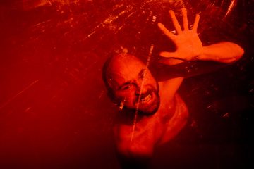 Kalju mies on veden alla, kurottaa toisella kädellään kohti pintaa ja hymyilee. Kuva kylpee kirkkaanpunaisessa valossa, ja veden aaltoilu ja heijastukset näkyvät hänen ympärillään.