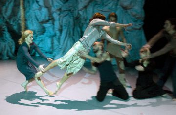 Ryhmä tanssijoita esiintyy lavalla; yksi valkoisiin pukeutunut henkilö hyppää vaakasuoraan, kun muut kurottautuvat, mikä luo liikkeen ja draaman tunnetta sinistä taustaa vasten.