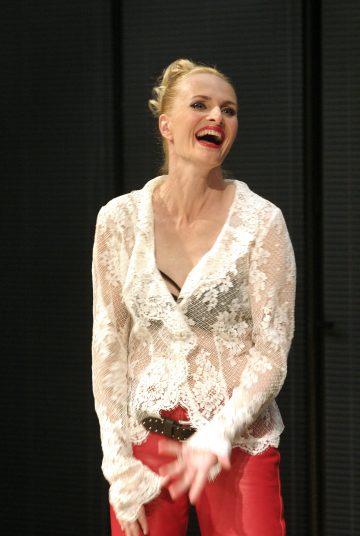 Nainen, jolla on vaaleat hiukset, hymyilee kirkkaasti. Hänellä on päällään läpikuultava valkoinen pitsinen pusero mustan topin päällä, punaiset housut ja musta vyö, ja hän seisoo tumman taustan edessä.