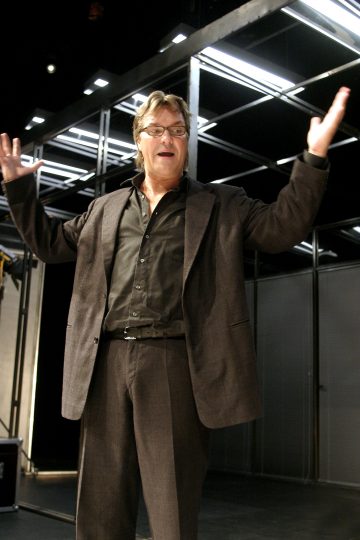 Silmälasipäinen ja tummaan pukuun pukeutunut mies seisoo sisätiloissa kädet kohotettuna ja ilmeikkäät kasvot modernin, geometrisen taustan edessä.