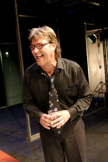 Mies, jolla on silmälasit, musta paita ja pilkullinen solmio, nauraa ja pitää kädessään juomaa hämärässä sisätilassa.