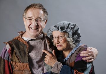 Vanha mies ja nainen, jotka ovat pukeutuneet tilkkutäkkimaisiin, vanhanaikaisiin vaatteisiin, hymyilevät toisilleen. Nainen, jolla on harmaa pipo, osoittaa leikkisästi miestä, jolla on silmälasit ja paikattu paita. He seisovat harmaan taustan edessä.