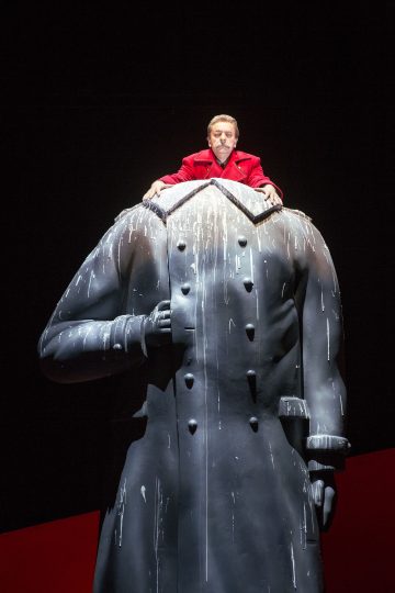 Punaiseen takkiin pukeutunut esiintyjä seisoo dramaattisella näyttämöllä suuren, päättömän, mustan patsaan takana ja yläpuolella, jolla on päällään maalattu takki, jonka pintaa pitkin kulkee valkoisia raitoja.