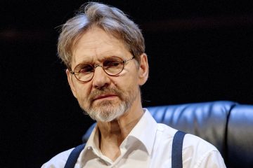 Keski-ikäinen mies, jolla on harmaat hiukset, parta ja pyöreät silmälasit, valkoinen paita ja tummat henkselit, istuu mustalla nahkatuolilla tummalla taustalla.