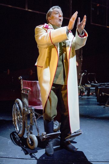 Pitkään kermanväriseen, punaisella koristeltuun takkiin pukeutunut vanhempi mies seisoo ja viittoo käsillään pyörätuolin edessä hämärästi valaistulla näyttämöllä, mikä viittaa teatteriesitykseen.