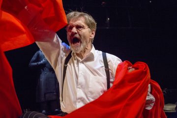 Vanhempi mies, jolla on silmälasit ja valkoinen paita, huutaa intohimoisesti pitäessään kädessään kirkkaanpunaista kangasta, ja taustalla on tumma tausta ja epäselvä hahmo kaukana.