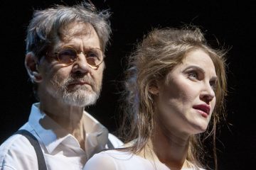 Iäkkäämpi mies, jolla on silmälasit ja valkoinen paita, seisoo nuoremman naisen takana, jolla on löysät hiukset ja punainen huulipuna, ja molemmat katsovat mietteliäästi kauas tummaa taustaa vasten.