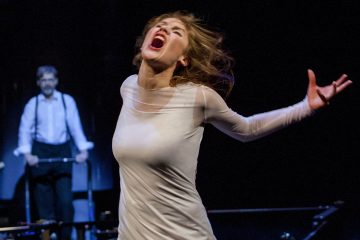 Valkoiseen mekkoon pukeutunut nainen huutaa intohimoisesti kädet ojennettuina tummalla näyttämöllä, kun taustalla seisoo mies, jolla on valkoinen paita ja henkselit, kävelijän vieressä.