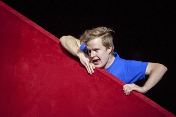 Siniseen paitaan pukeutunut nuori henkilö nojaa suuren, vino punaisen pinnan yli ja vaikuttaa eloisalta ja ilmeikkäältä tummaa taustaa vasten.