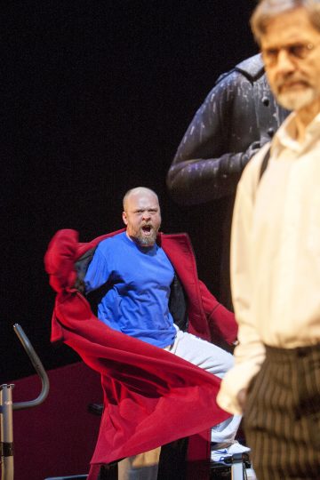 Siniseen paitaan ja punaiseen takkiin pukeutunut mies seisoo keskellä toimintaa suu auki huutaen, kun taas toinen mies, jolla on silmälasit ja valkoinen paita, seisoo etualalla, epätarkkana. Kohtaus vaikuttaa dramaattiselta ja teatraaliselta.