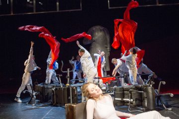 Harmaisiin pukuihin pukeutuneet näyttelijät heiluttelevat energisesti suuria punaisia kangaskappaleita sotkuisella näyttämöllä, kun taas valkoisiin pukeutunut nainen istuu etualalla silmät kiinni ja luo dramaattisen ja dynaamisen teatterikohtauksen.