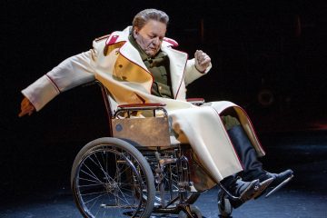 Henkilö, jolla on pitkä valkoinen takki, jossa on punainen reunus ja tummat saappaat, istuu pyörätuolissa hämärästi valaistulla näyttämöllä ja elehtii dramaattisesti toisella kädellään.