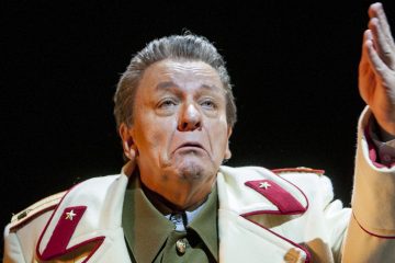 Vanhempi mies, jolla on sotilaspuku, jossa on punaisia ja kultaisia aksentteja, elehtii oikealla kädellään, ja hänen kasvoillaan näkyy dramaattinen tai ilmeikäs tunne tummaa taustaa vasten.