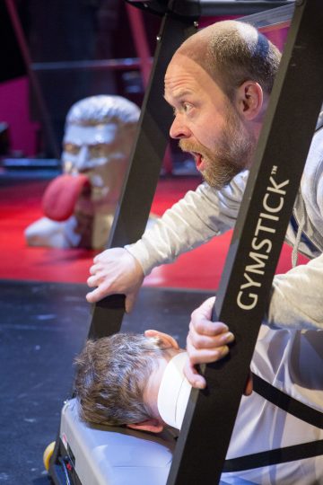 Mies tarttuu juoksumattoon, jossa lukee GYMSTICK, ja näyttää järkyttyneeltä, kun toinen henkilö makaa kasvot alaspäin juoksumatolla. Taustalla näkyy suuri nyrkkeilynukke, jolla on punainen hanska.