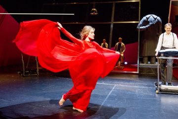 Paljasjalkainen nainen virtavassa punaisessa mekossa tanssii energisesti lavalla, kangas hulmuaa hänen ympärillään. Taustalla seisoo kolme miestä, yksi näppäimistön ääressä, kaikki modernissa teatteriympäristössä, jossa on dramaattinen valaistus.