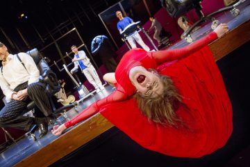 Nainen punaisessa mekossaan nojaa dramaattisesti taaksepäin lavan reunan yli, suu auki kuin huutaen. Hänen takanaan useat ihmiset istuvat ja seisovat toimistotuolien ääressä teatterikohtauksessa.