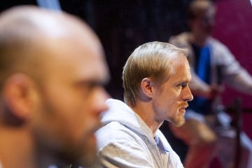 Vaaleat hiukset ja partainen mies, jolla on vaalea huppari, on tarkennettuna ja katsoo oikealle. Toinen mies etualalla ja henkilö taustalla ovat molemmat epätarkkoja.