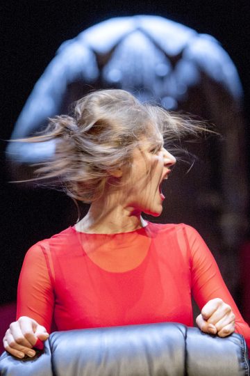 Läpikuultavaan punaiseen toppiin pukeutunut nainen puristuu tuoliin ja huutaa, ja hänen hiuksensa lentävät villisti sivulle. Tausta on epätarkka, ja siinä on tumma, koristeellinen rakennelma, joka muistuttaa siipiä tai valtaistuinta.