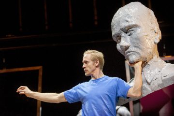 Lavalla seisoo sinipukuinen mies, joka pitää suurta harmaata veistettyä päätä kaulasta ja ojentaa toisen kätensä ulospäin. Tausta on tumma, mikä korostaa dramaattista kohtausta.