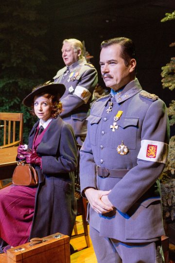 Lavalla on kolme ihmistä 1900-luvun alun sotilas- ja aikalaispuvuissa. Harmaapukuinen mies seisoo edessä, kun taas hattuun ja takkiin pukeutunut nainen istuu pöydän ääressä, ja toinen univormuun pukeutunut mies seisoo hänen takanaan.
