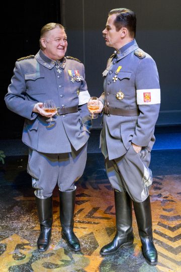 Kaksi miestä harmaissa sotilaspuvuissa ja mitaleissa seisoo vierekkäin, ja kummallakin on juoma lasissa. He näyttävät keskustelevan lavalla kirkkaassa valaistuksessa.