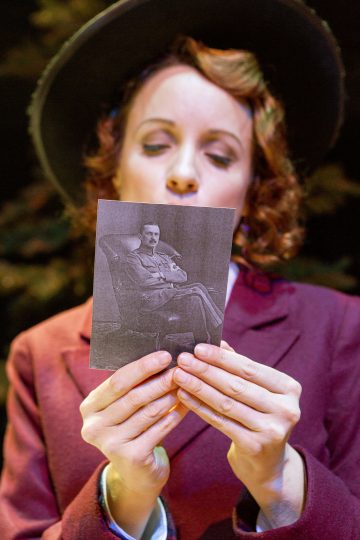 Nainen, jolla on ruskeankeltainen takki ja leveälakinen hattu, pitää kädessään mustavalkoista valokuvaa tuolilla istuvasta miehestä, joka katsoo kuvaa mietteliäästi.