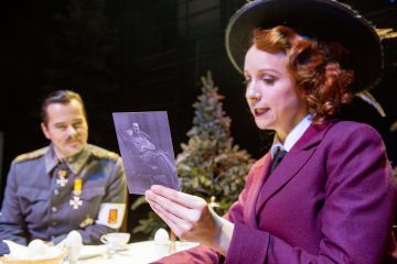 Nainen, jolla on vanha violetti asu ja hattu, pitelee mustavalkoista valokuvaa istuessaan pöydän ääressä sotilaspukuun pukeutuneen miehen kanssa, ja molemmat keskustelevat, ja taustalla näkyy joulukuusia.