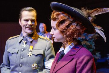 Mies, jolla on koristeltu sotilasunivormu, hymyilee punaiset kiharat hiukset omaavalle naiselle, jolla on leveälippainen hattu, jossa on höyheniä, ja ruskeankeltainen takki, ja molemmat esiintyvät teatterilavalla.