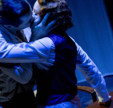 Kaksi ihmistä syleilee ja suutelee intohimoisesti näyttämöllä dramaattisessa valaistuksessa, toinen käsi toisen kasvoilla, molemmilla on aikakauden tyyliset puvut.