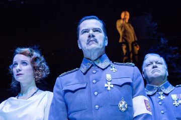 Kolme sotilaspukuista näyttelijää seisoo kirkkaiden näyttämövalojen alla vakavana. Vasemmalla seisoo valkopukuinen nainen, hänen vieressään on kaksi sinisiin univormuihin pukeutunutta miestä, ja heidän yläpuolellaan taustalla seisoo varjoinen hahmo.