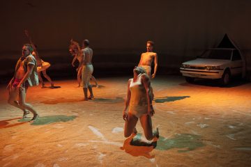 Kuusi vähäpukeista tanssijaa tekee ilmeikkäitä liikkeitä hiekalla päällystetyllä näyttämöllä, jossa on dramaattinen valaistus ja oikealla puolella näyttämöä pysäköity valkoinen auto, joka on osittain peittynyt tumman taustan alle.