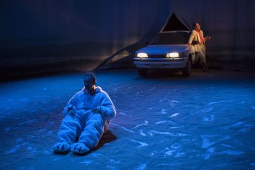 Valkoiseen karhupukuun pukeutunut henkilö istuu hämärästi valaistulla hiekkapohjaisella näyttämöllä, kun taas toinen henkilö seisoo taustalla valkoisen auton vieressä pitelemässä jotakin sinisten ja vaaleanpunaisten valojen valaisemana.