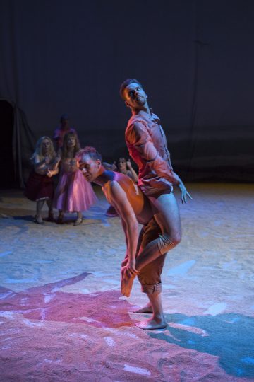 Kaksi miestanssijaa esittää ilmeikkään ohjelman hämärästi valaistulla näyttämöllä, kun joukko vaaleanpunaisiin pukuihin pukeutuneita ihmisiä katselee taustalla. Lattia näyttää hiekkaiselta, ja värikkäät näyttämövalot heittävät dramaattisia varjoja.