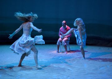 Kaksi naista virtaavissa mekoissa tanssii energisesti hiekkapohjaisella lavalla sinisessä valossa, kun vaaleanpunaiseen paitaan ja shortseihin pukeutunut mies istuu katsomassa valkoiselta tuolilta taustalla.