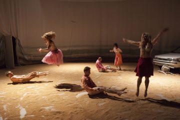 Kuusi ihmistä poseeraa ja hyppii hiekkatäytteisellä lattialla lämpimässä valossa; kaksi naista on ilmassa hameissaan, kun taas muut istuvat tai lepäävät. Valkoinen auto on pysäköity oikealle, tuolin lähelle. Kohtauksessa on surrealistinen, performanssimainen tunnelma.