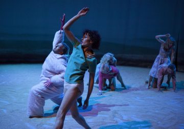 Viisi esiintyjää on hiekkapohjaisella näyttämöllä dramaattisissa asennoissa. Yhdellä on yllään pörröinen valkoinen puku, kun taas muilla on rennot tai pastelliväriset vaatteet. Sininen valaistus luo teatraalisen, surrealistisen tunnelman.